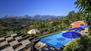 A pool area with lounge chairs and umbrellas, surrounded by lush greenery and mountains in the background. - Relaxing Retreat in Le Marche (Monte San Martino)