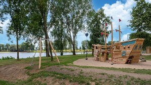 Een speeltuin met een piratenschip, schommels en een zandbak, gelegen bij een meer.