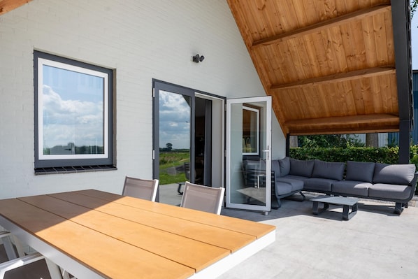 Une terrasse extérieure avec une table et des chaises en bois, un canapé gris et une porte vitrée donnant sur un jardin.