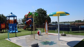 Ein Spielplatz mit Rutschen, einer Kletterkonstruktion und einem Wasserspielbereich mit einem großen Sonnenschirm.