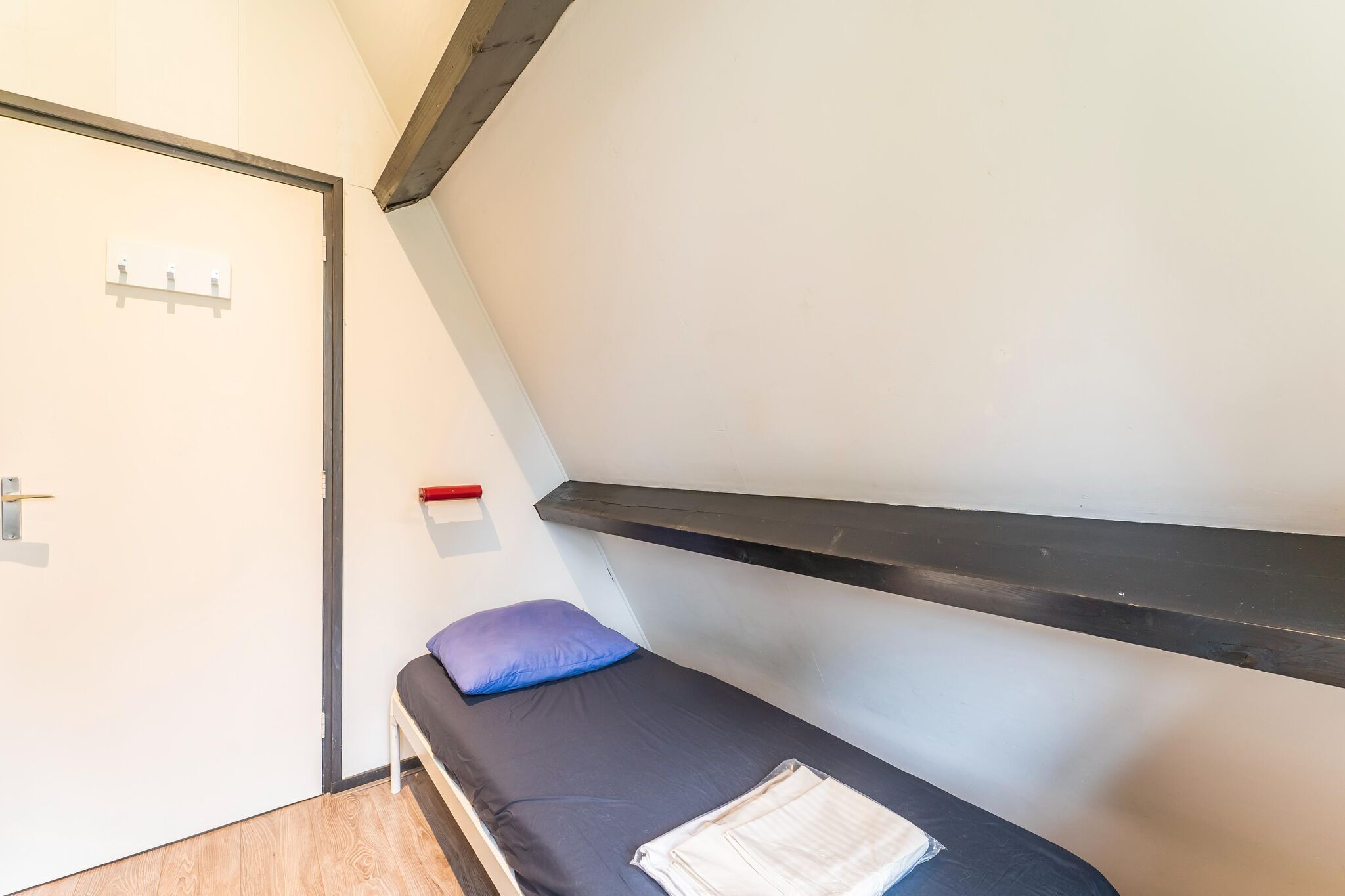 A small, single-bed room with a dark mattress, a blue pillow, and a white blanket on the bed. There is a white door with a handle and a light switch on the wall.