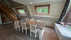 Een eetruimte met een houten tafel en witte stoelen, een trap en een raam met uitzicht op bomen.