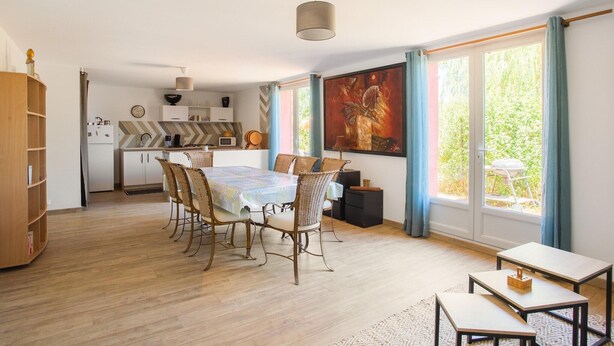 A dining area with a table and chairs, a kitchen in the background, and a large window with curtains.