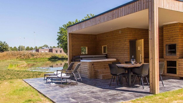 Eine moderne Holzblockhütte mit einer Terrassenfläche, die einen Tisch, Stühle, einen eingebauten Grill und Blick auf ein Gewässer bietet.