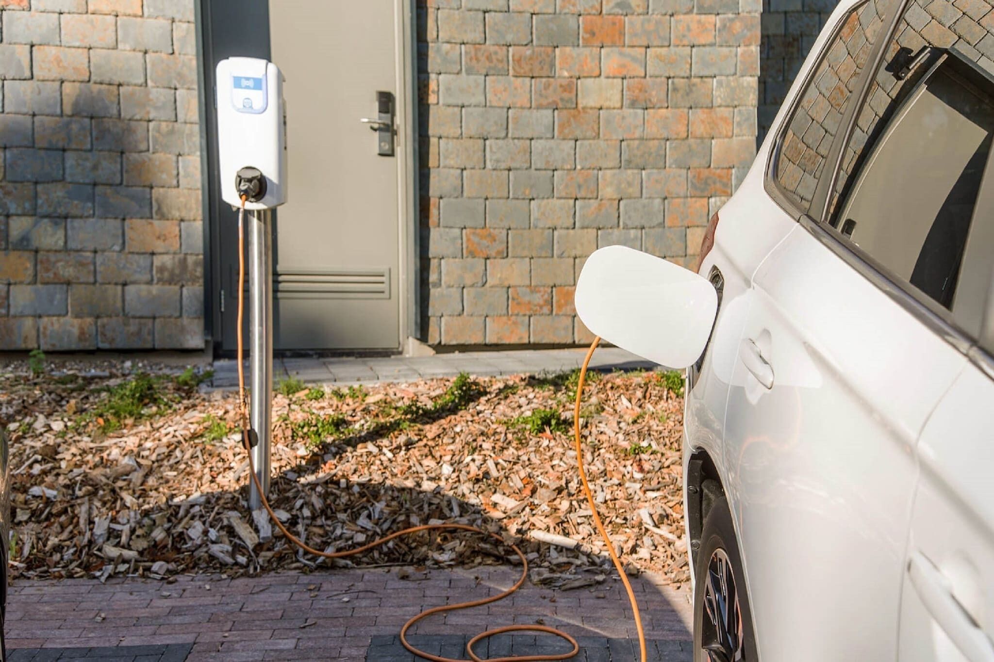 Une voiture blanche est en train de se recharger à une borne de recharge publique, avec un mur en briques et une porte en arrière-plan.
