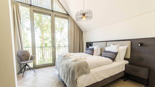 A bedroom with a large bed, a chair, a nightstand, and a view of greenery through the window.