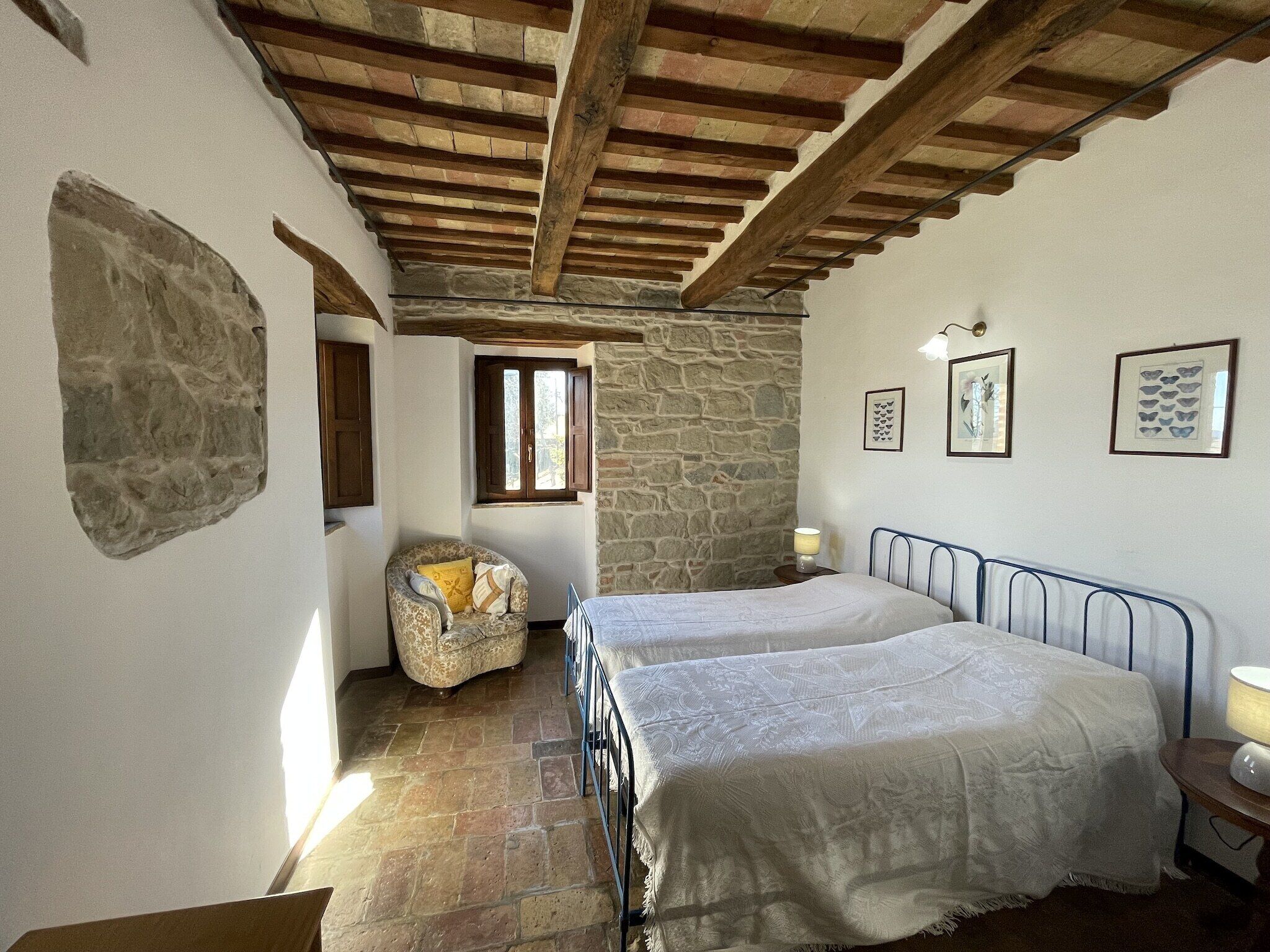 Un dormitorio con pared de piedra, cama con estructura metálica, una silla y una mesita de noche con lámpara.