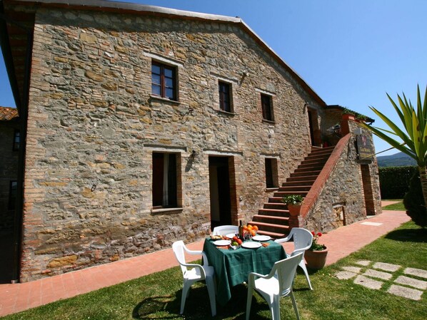 Un edificio de piedra con una escalera que conduce a un segundo piso. Hay una mesa con sillas y un mantel colocado en el exterior.