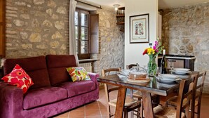 Una habitación acogedora con pared de piedra, una mesa de comedor de madera dispuesta con platos y un florero, y un sofá rojo suave con cojines de colores.