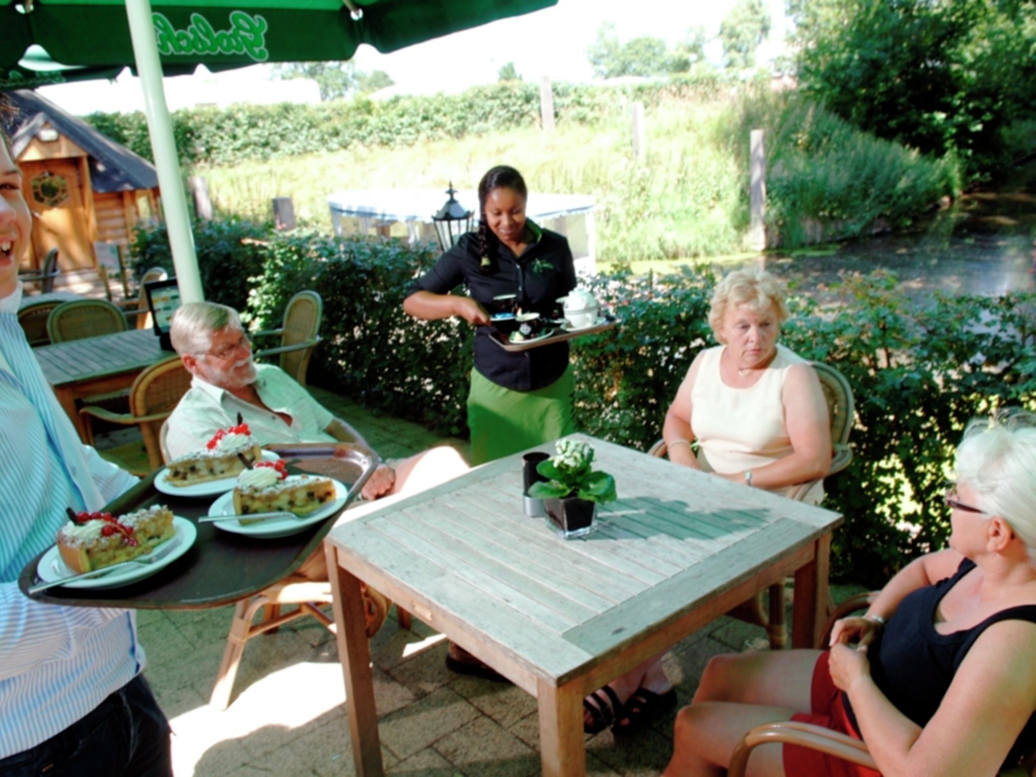 A group of people seated at an outdoor table with a menu, surrounded by greenery and a pond.