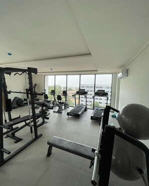 A gym with exercise equipment, a bench, and a view of buildings through large windows. - Park Apartments D-plus One bed+Living room - Cantonments  (Accra)
