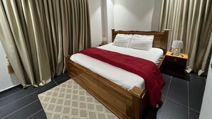 Une chambre d’hôtel avec un lit en bois, des rideaux beiges, une table de chevet avec une lampe et un tapis à motifs.