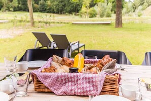 Ein Picknick mit Korb, darin Brot, Eier und eine Orangensaftflasche auf einem Tisch, mit Stühlen und einer Wiese im Hintergrund.