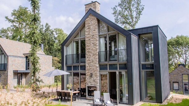 Une maison moderne avec une façade en pierre, de grandes fenêtres et un patio aménagé avec du mobilier d’extérieur.