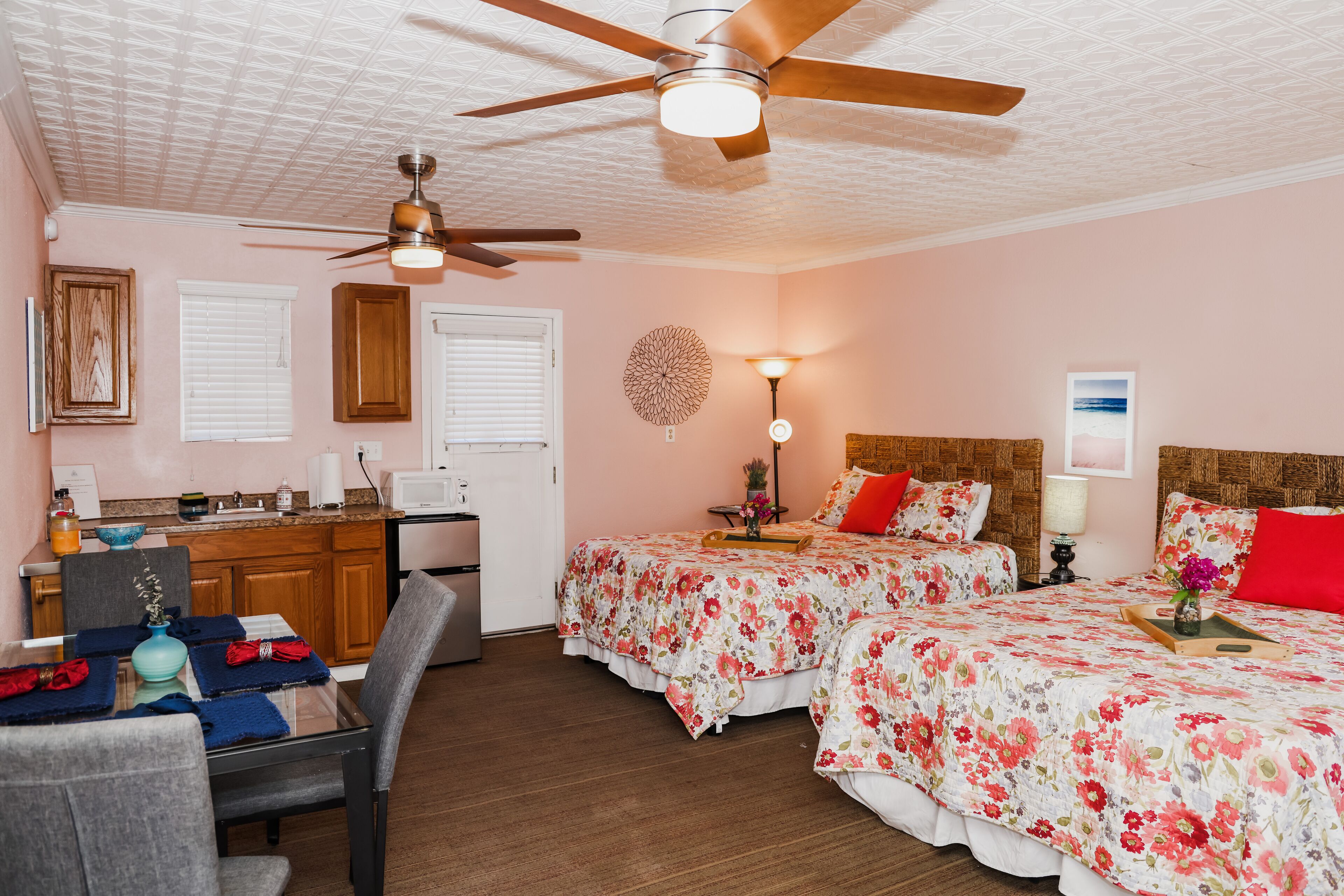 Quarto com duas camas, um ventilador de teto, uma escrivaninha e um espaço de jantar com cadeiras e mesa.
