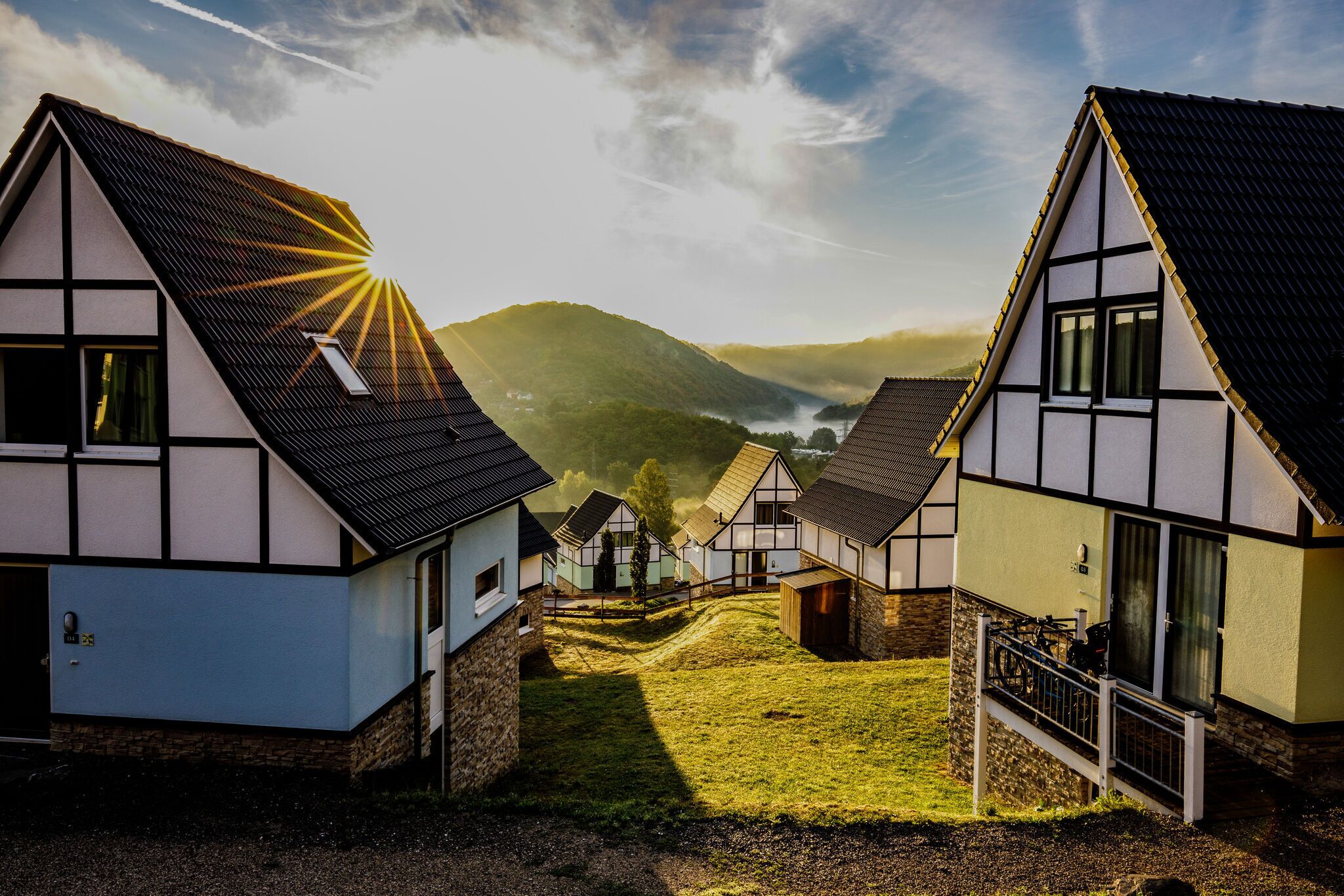 A row of traditional half-timbered houses with sloped roofs, a grassy pathway, and a mountainous backdrop.