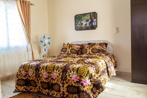 Un lit bien fait, recouvert d’une couvre-lit à motifs, un ventilateur fixé au mur et un tableau représentant une cascade.