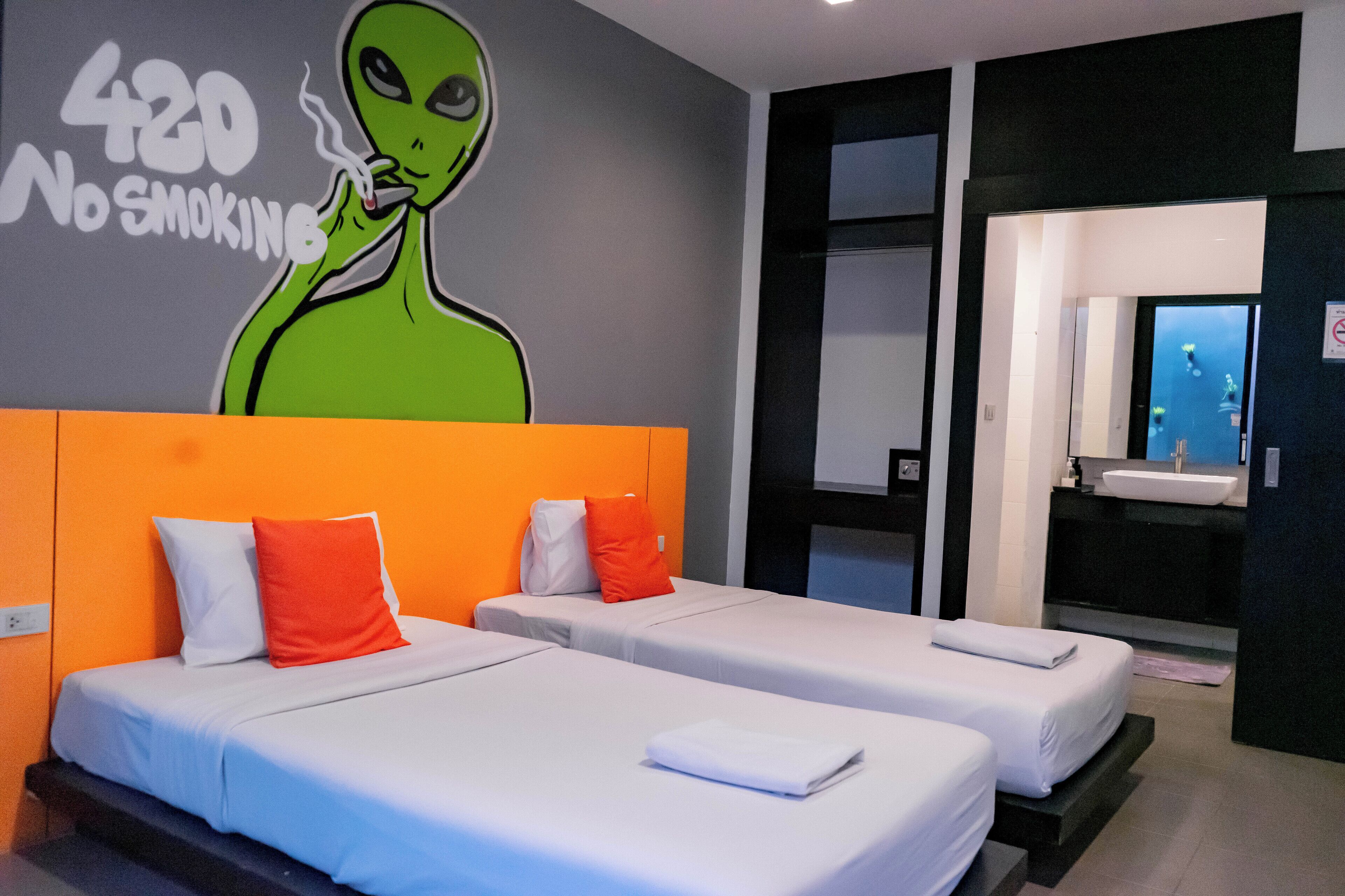 A hotel room with two beds, a mural of an alien smoking, and a bathroom with a sink and mirror.