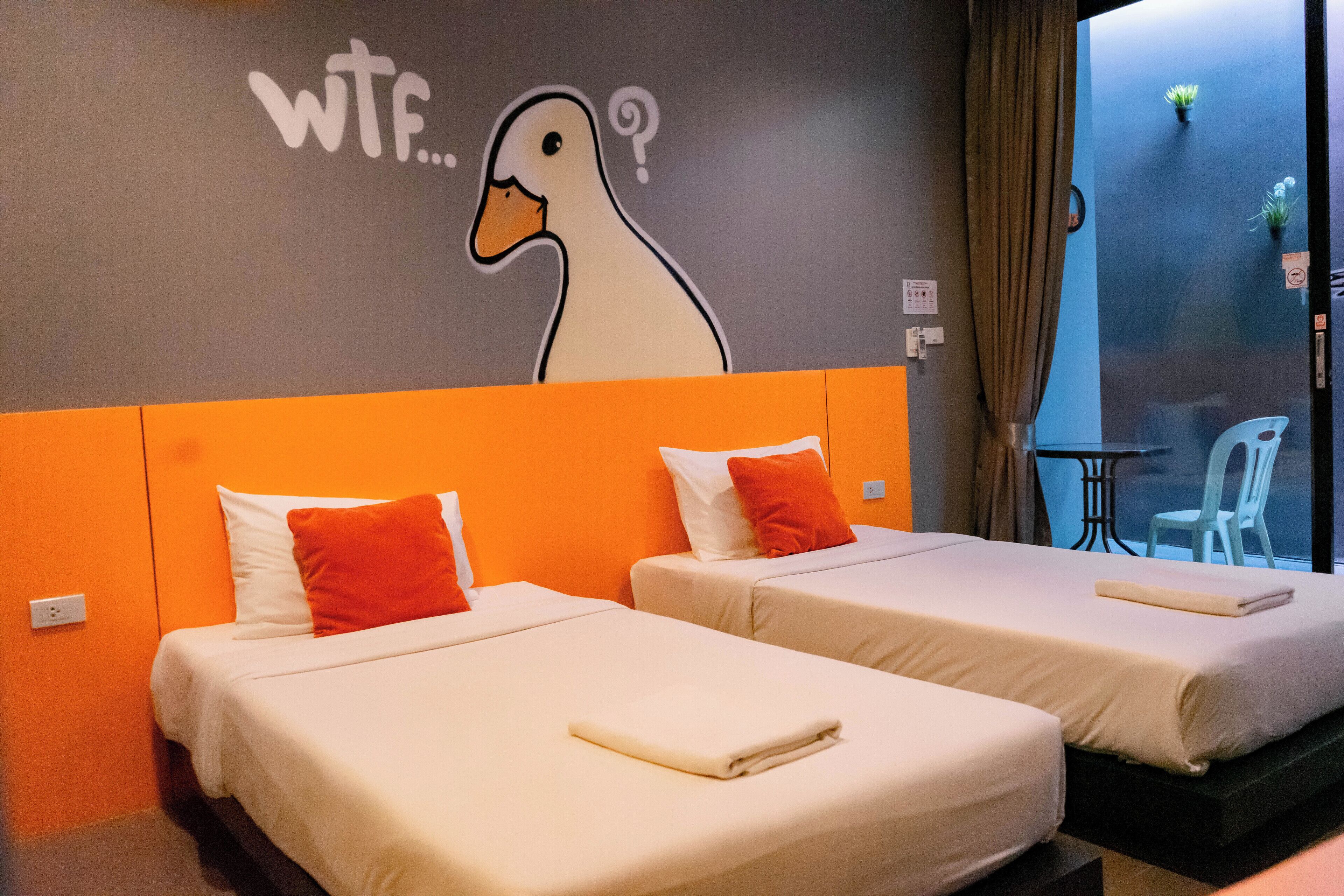Deux lits avec une literie blanche et orange, une fresque murale représentant un canard, et une vue sur un balcon avec une chaise et des plantes.