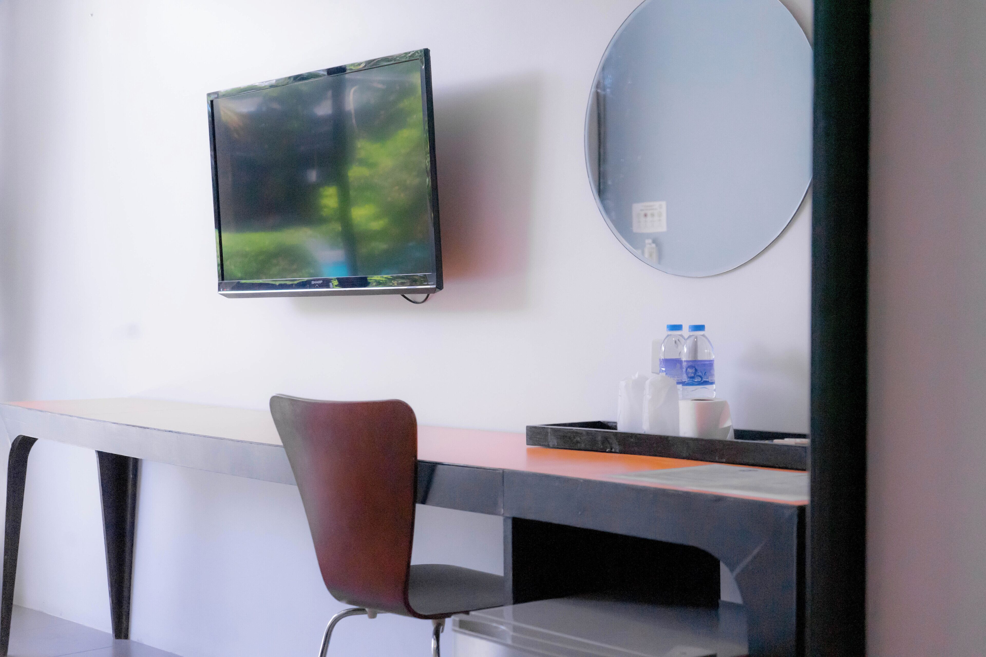 A hotel room with a flat-screen TV mounted on the wall, a round mirror, a desk with a chair, and a water bottle on the desk.