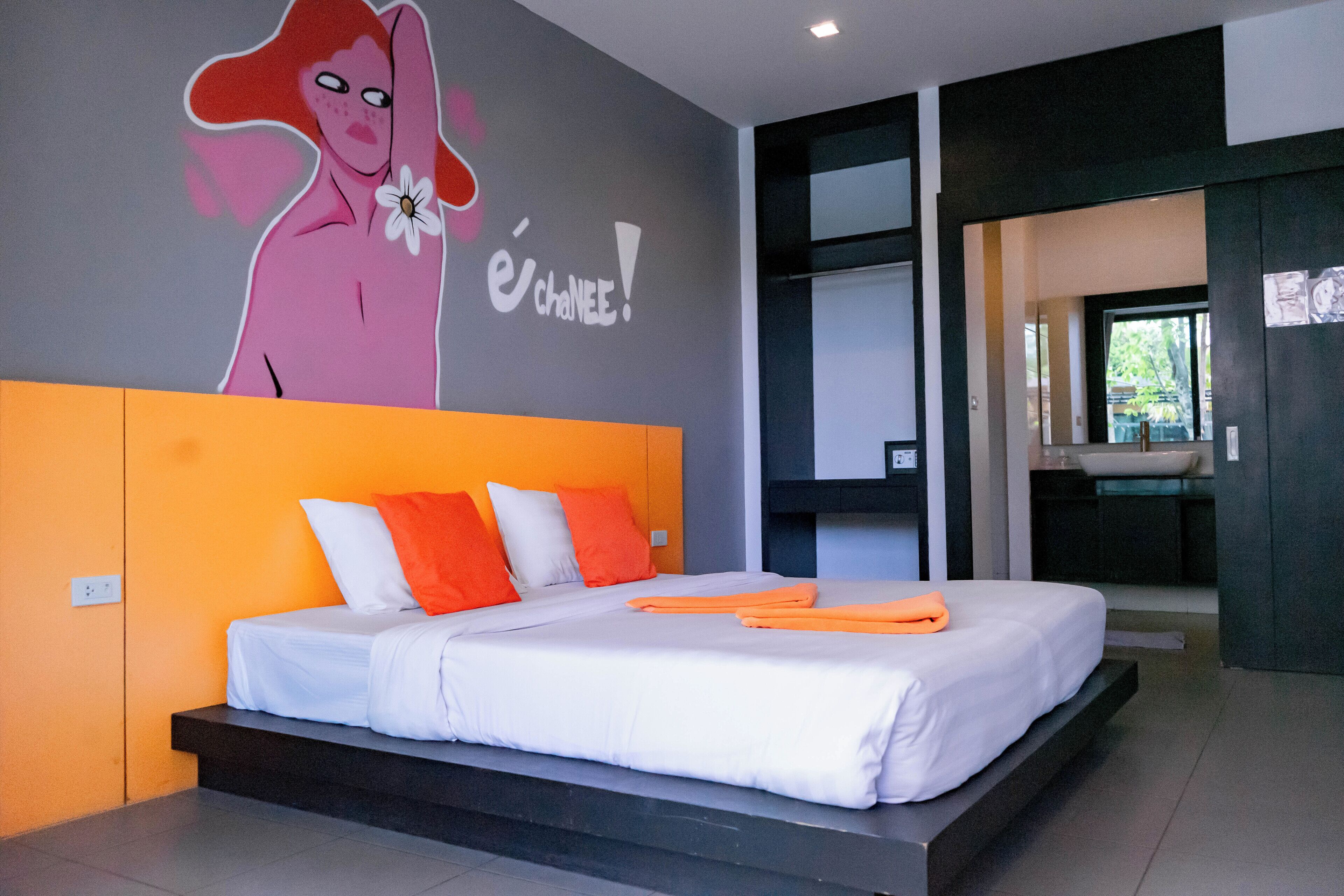 A modern bedroom with a large bed, orange and white bedding, a mural on the wall, and a bathroom visible through an open door.