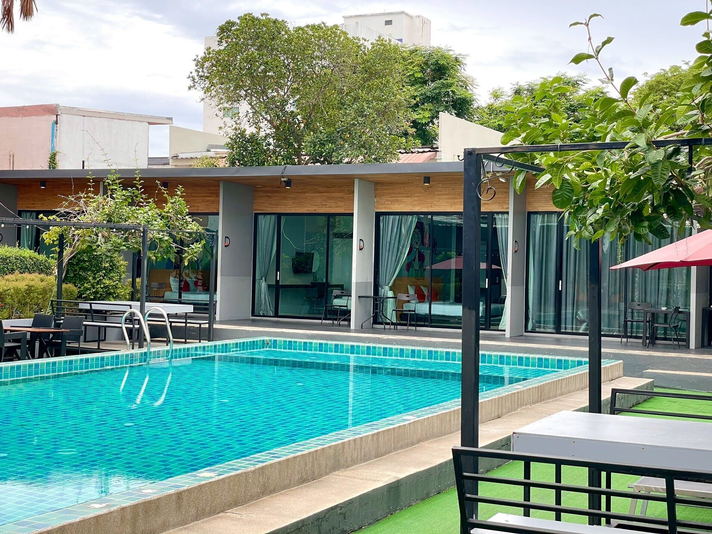 Une piscine dont le bord est revêtu de carrelage, entourée d’un espace extérieur aménagé avec des sièges et d’une zone couverte avec un parasol rouge.