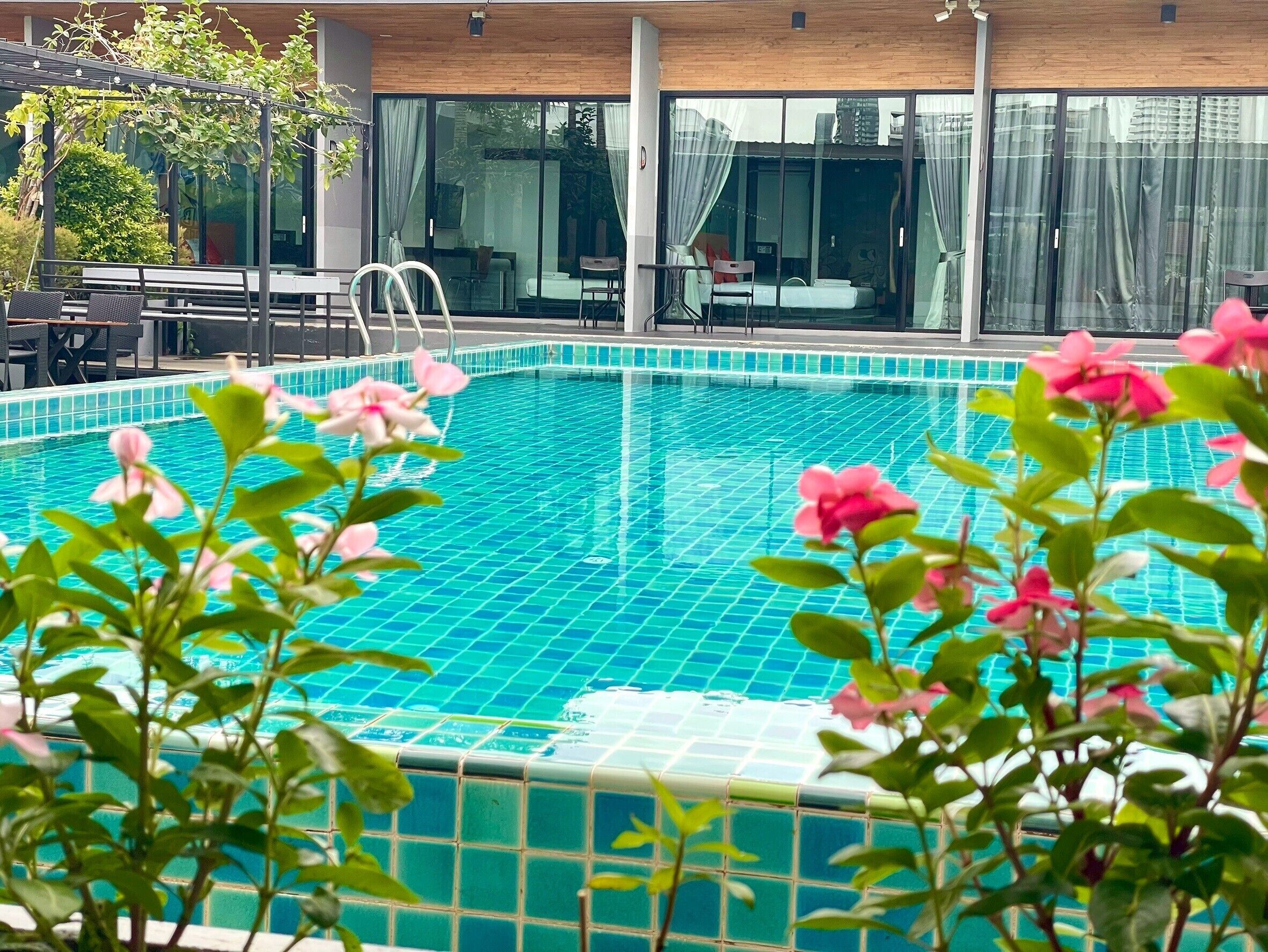A swimming pool with a tiled edge, a covered seating area with tables and chairs, and a building with large windows and curtains.