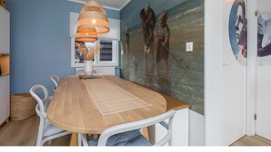 A dining area with a wooden table, white chairs, a woven pendant light, and a mural of children on a beach.
