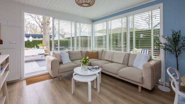 A modern living room with a beige sectional sofa, a white coffee table, and a large window with white blinds.