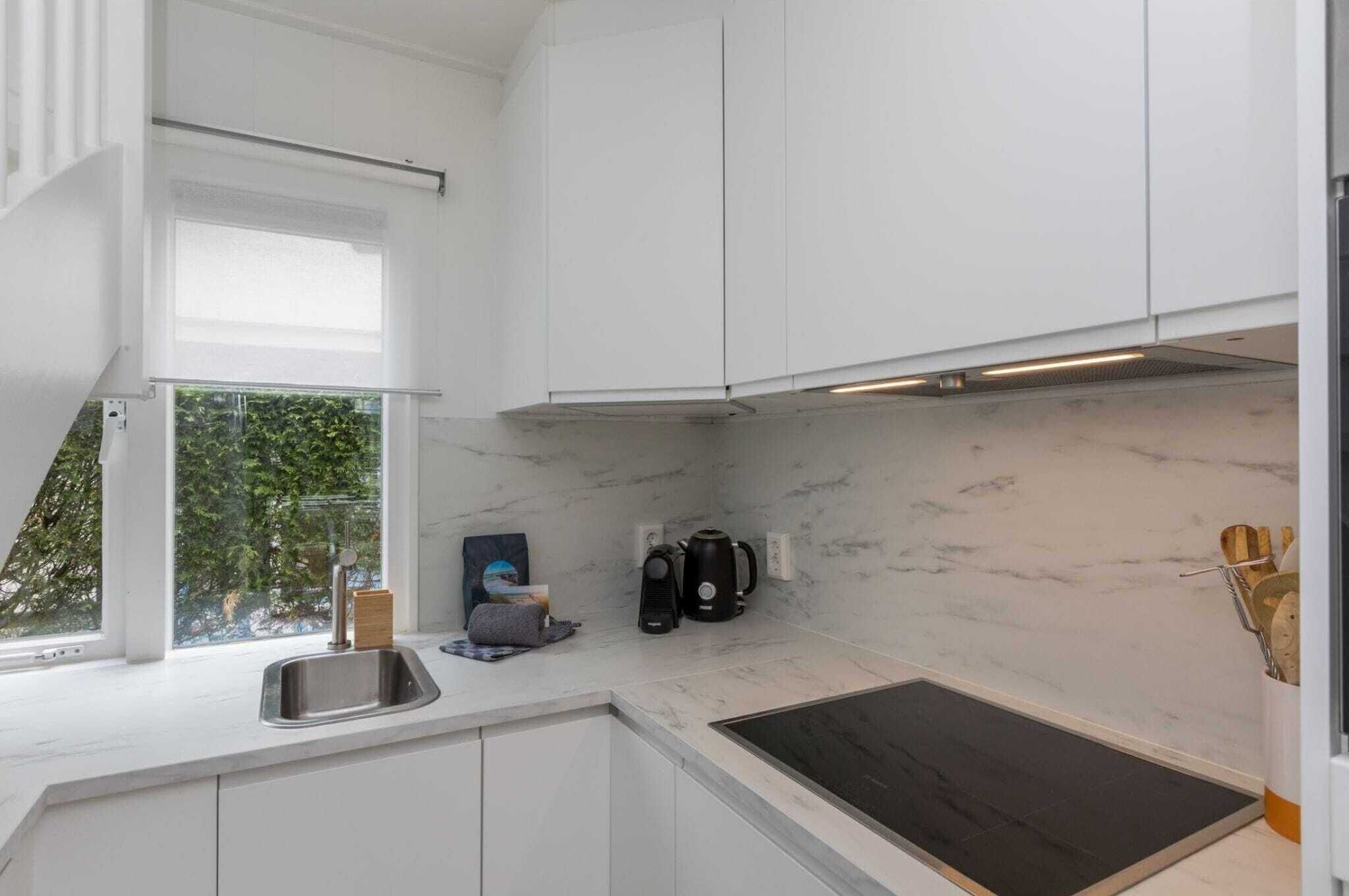 Une cuisine moderne avec des armoires blanches, un pare-feu en marbre, une cuisinière intégrée et une fenêtre donnant sur un espace verdoyant.