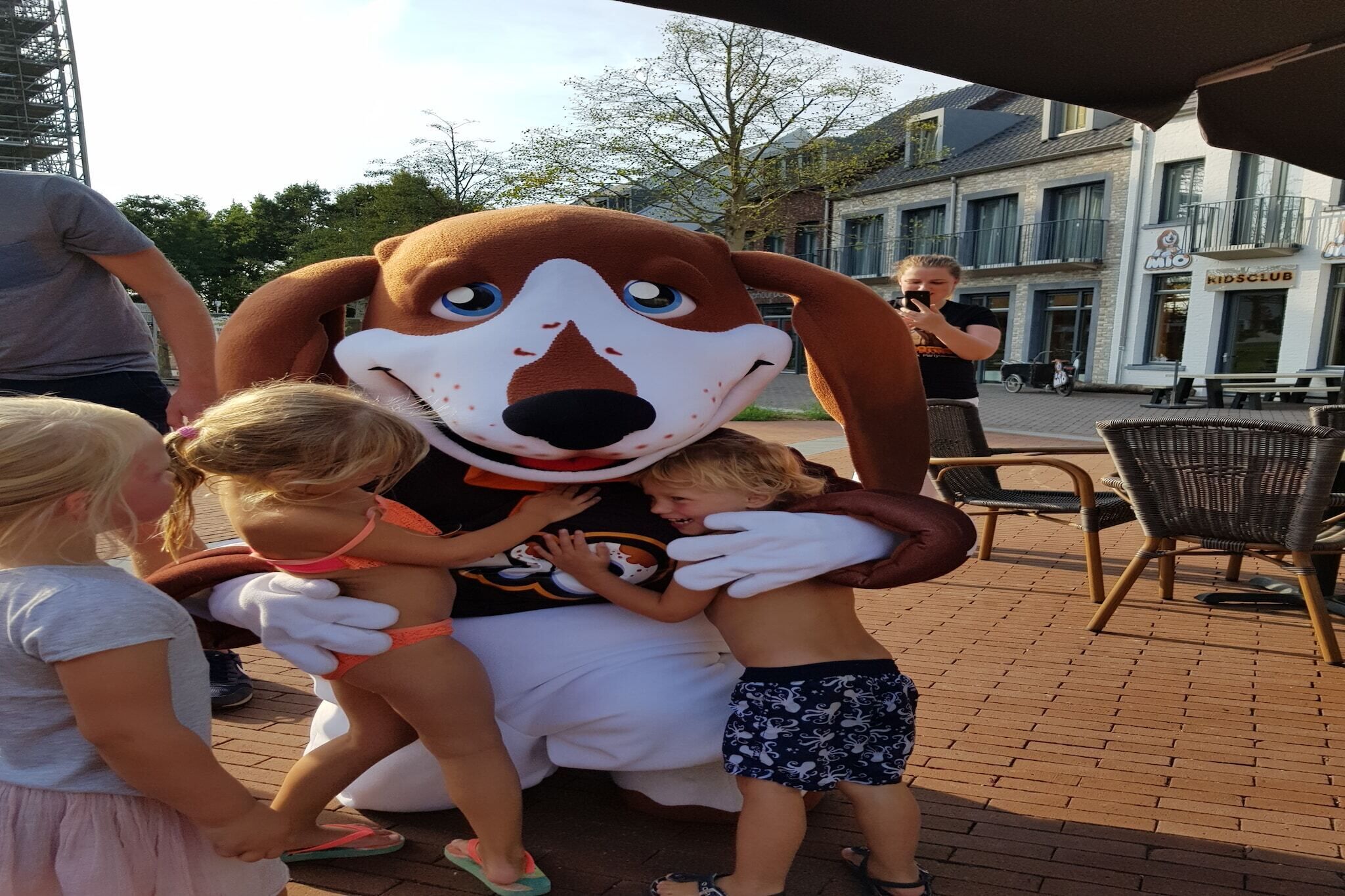 A mascot interacting with children on a paved outdoor area with chairs and tables, and a building with a sign that reads "KIDS CLUB" in the background.