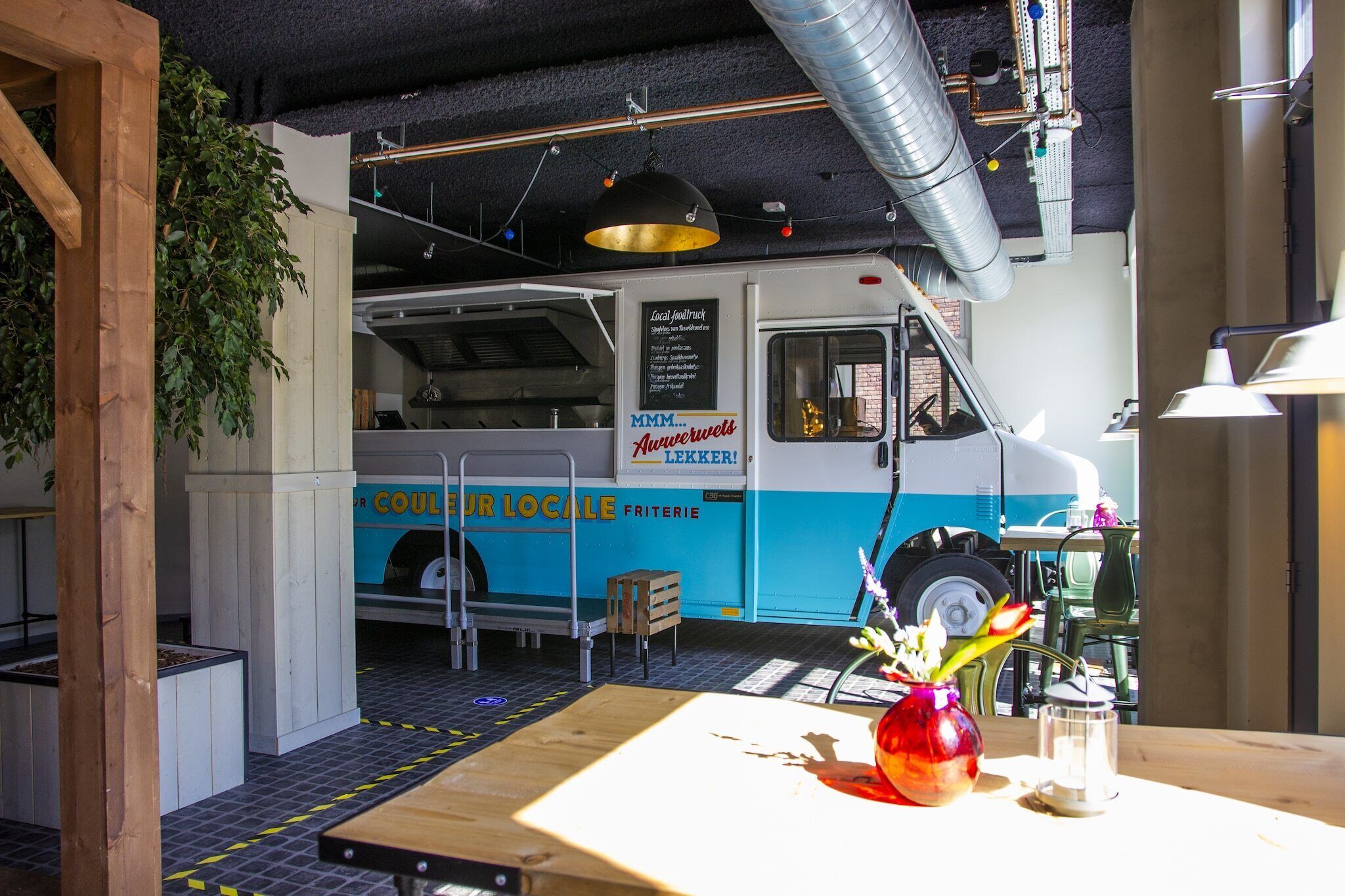 A food truck with a menu board, a wooden table with a vase, and a hanging light fixture.