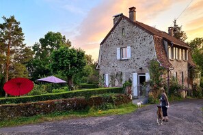 Una casa de piedra con techo inclinado, un camino de entrada pavimentado y un perro cerca de la entrada.