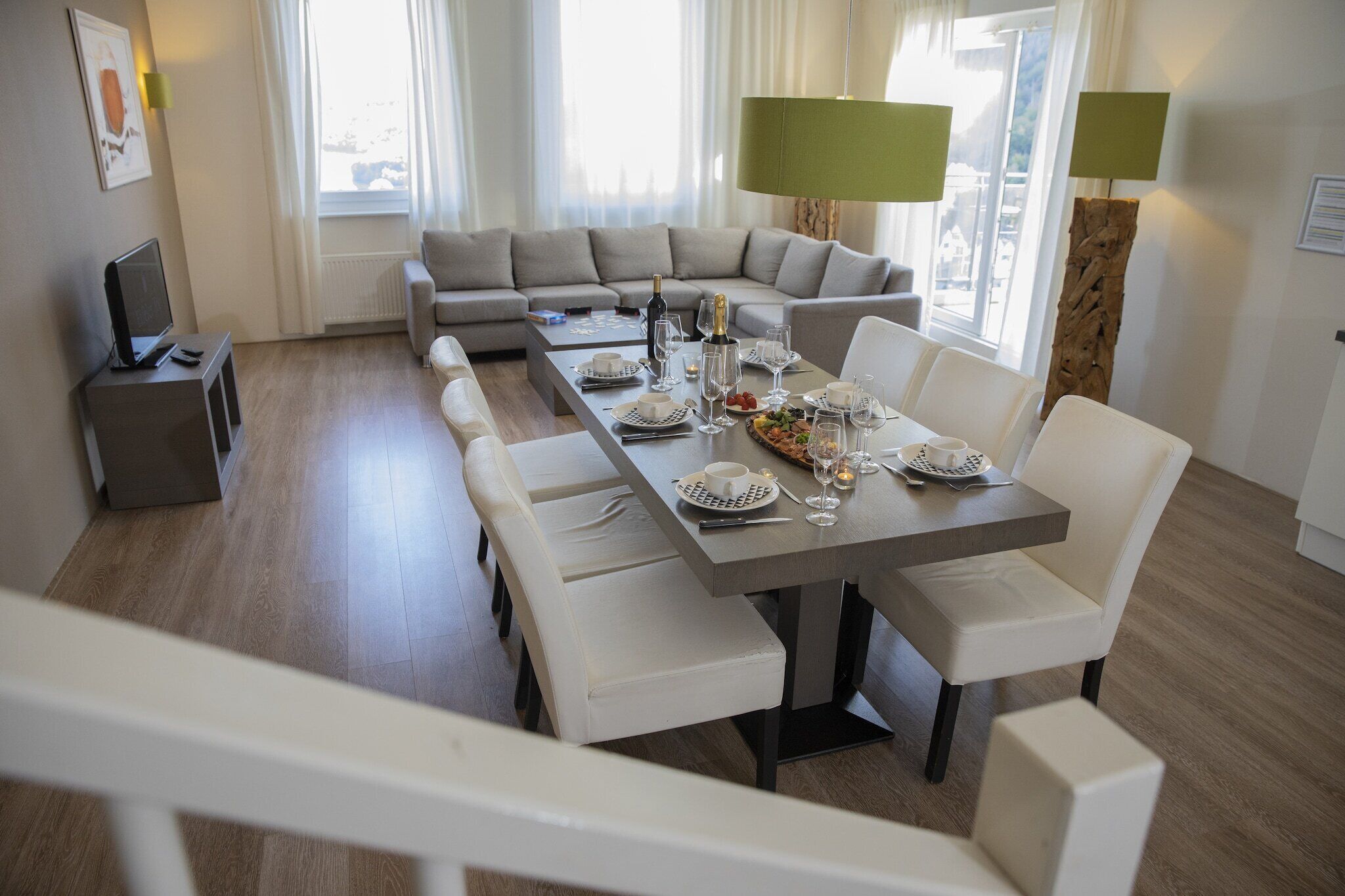 A modern dining area with a long table set for six, a sofa, and a TV in a bright room with large windows.