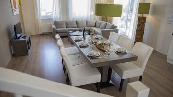 Un espace repas moderne avec une longue table dressée pour six personnes, un canapé et un téléviseur, le tout dans une pièce lumineuse dotée de grandes fenêtres.