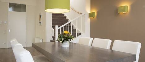 Un espace repas avec une longue table, des chaises blanches, un vase de fleurs, un escalier et des luminaires fixés au mur.