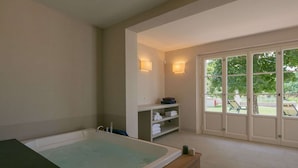 A spa room with a large bathtub, shelves with towels, and a view of a garden through a glass door.