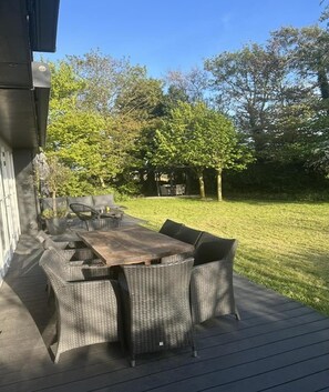 Eine Terrasse im Freien mit einem Holztisch und Korbstühlen, umgeben von einer Rasenfläche und Bäumen.