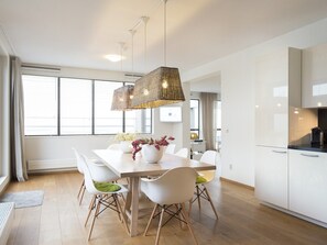 A modern kitchen with white cabinets, a dining area with a wooden table and white chairs, and a large window with blinds.