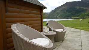 A wooden cabin with wicker chairs and a table with a bottle and glasses, overlooking a lake and mountains.