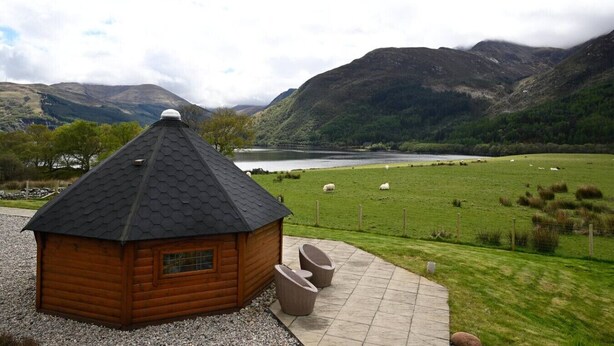 A wooden cabin with a sloped roof, a small window, and a door, situated on a gravel path with a paved area in front.