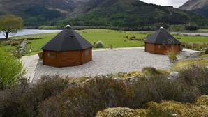 Two wooden cabins with sloped roofs, surrounded by a gravel path, a grassy field, and mountains in the background. - Leven View Lodges near Glencoe No2 Callart (Fort William)