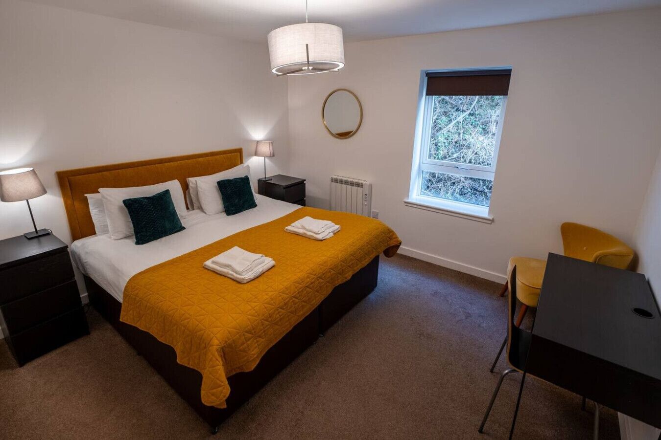 A bedroom with a bed, two bedside tables, a desk, a chair, a window, and a radiator.