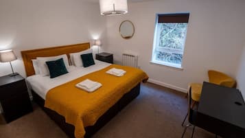 A bedroom with a bed, two bedside tables, a desk, a chair, a window, and a radiator.