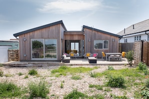 A modern two-story house with a large sliding glass door entrance, a patio with outdoor seating, and a garden area with gravel and plants.