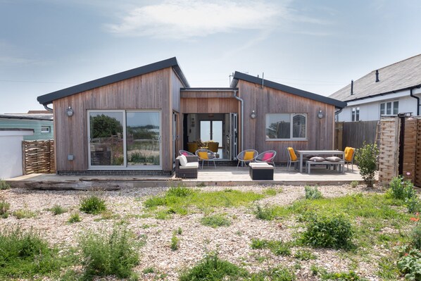 Une maison moderne à deux étages, dotée d’une grande porte-fenêtre coulissante à l’entrée, d’un patio avec coin salon extérieur et d’un jardin agrémenté de gravier et de plantes.