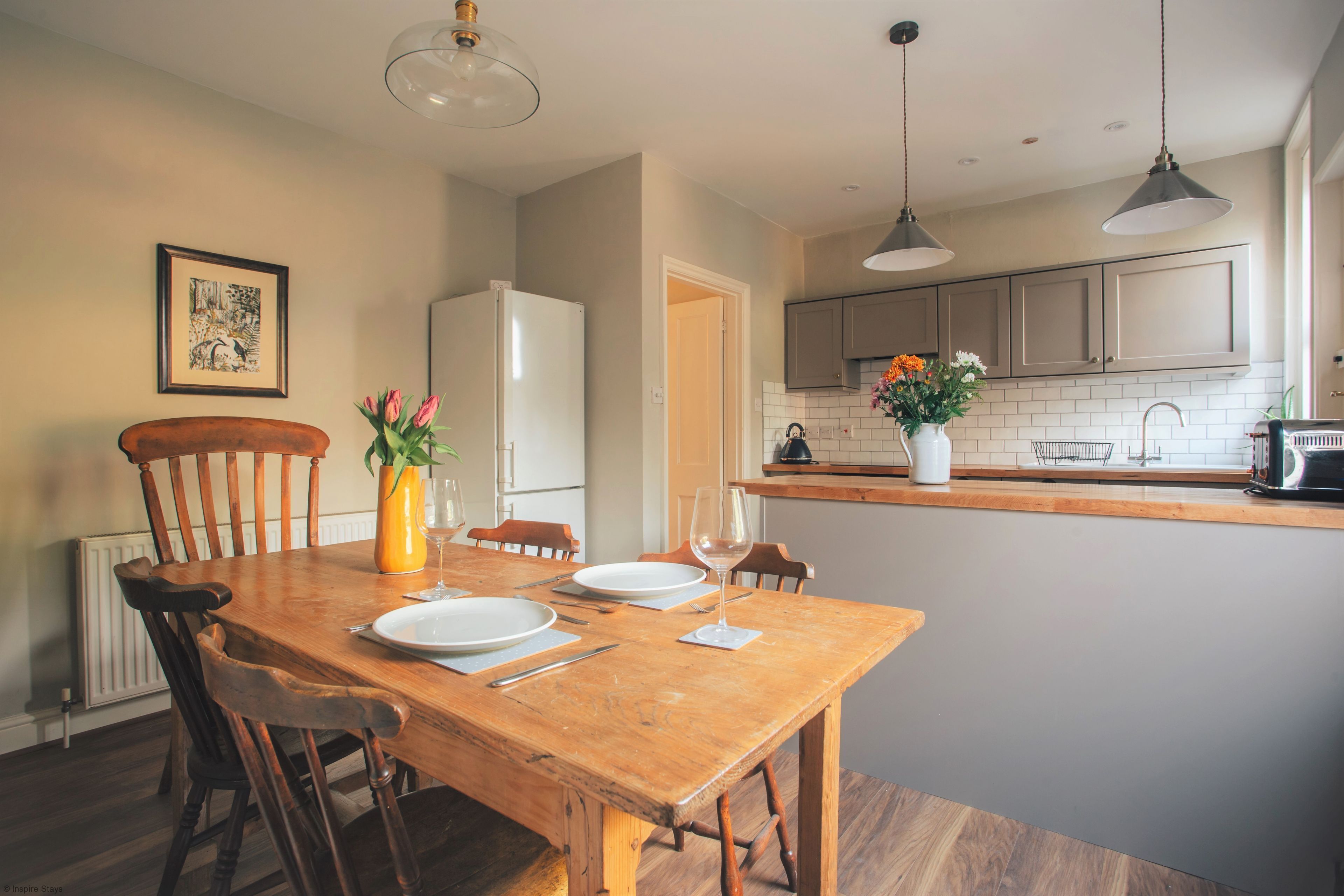 Un comedor con una mesa de madera dispuesta para dos personas, un refrigerador y una cocina con mostrador y armarios.