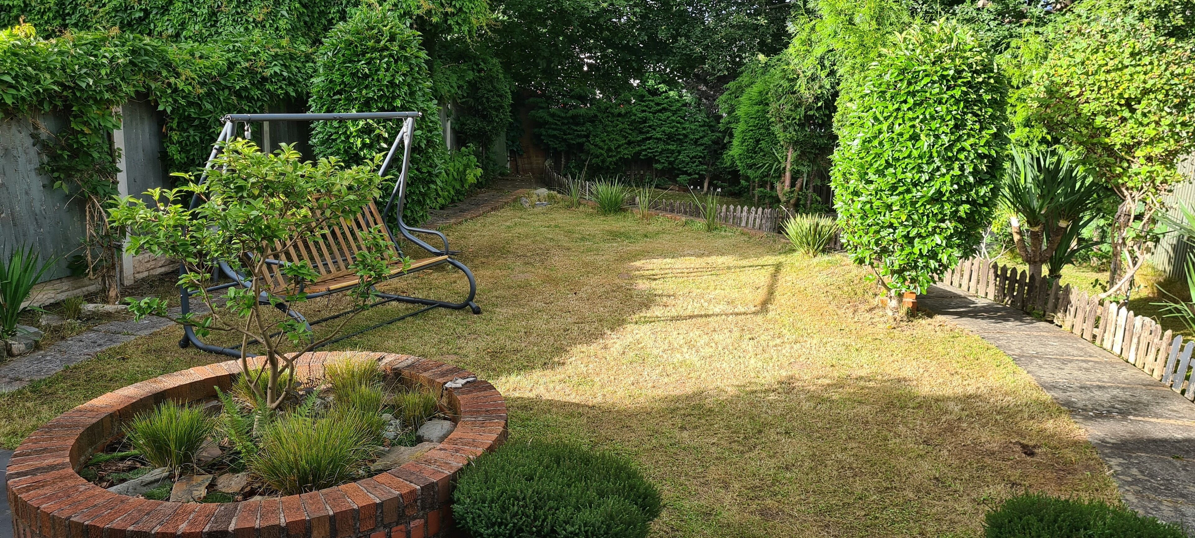 A garden with a swing set, a brick planter with plants, and a wooden fence.