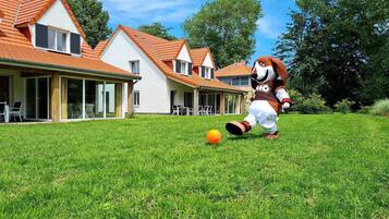 Une mascotte se trouve dans un espace verdoyant, avec une maison en arrière-plan.