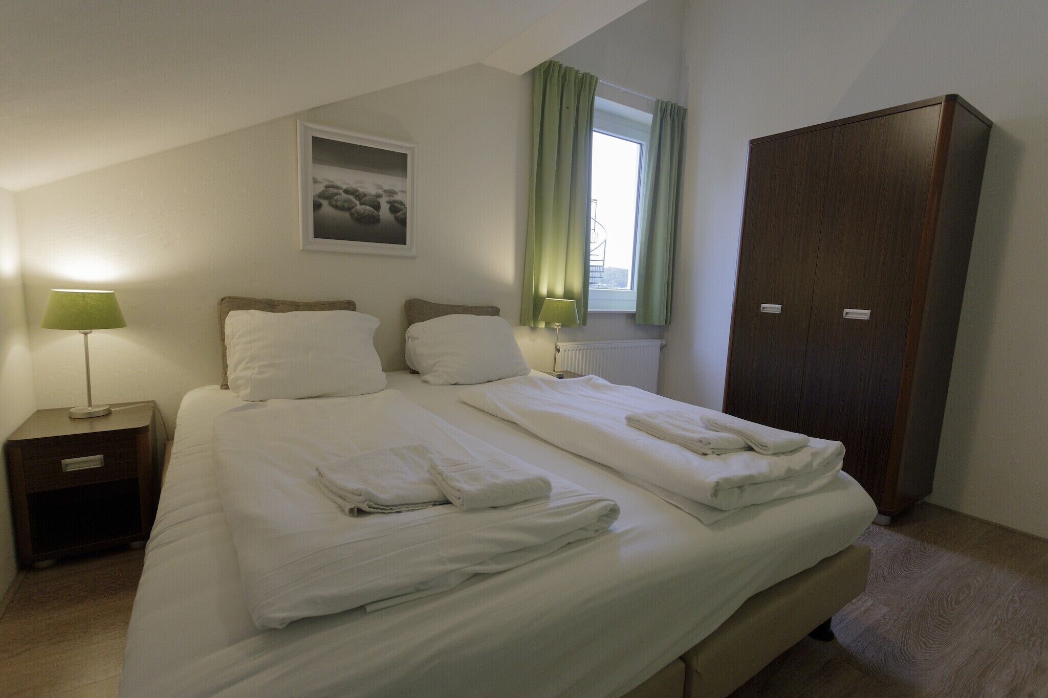 Un lit jumeau bien fait, avec du linge de lit blanc, une table de chevet avec une lampe, une armoire et une fenêtre avec des rideaux.